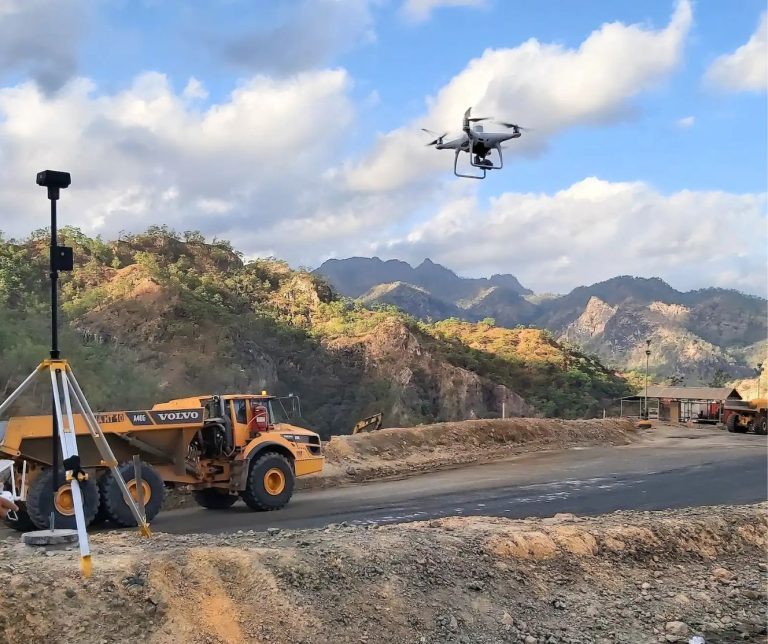 Drone conducting an aerial survey over an active mining construction site, with a surveying instrument and articulated dump truck in the foreground.